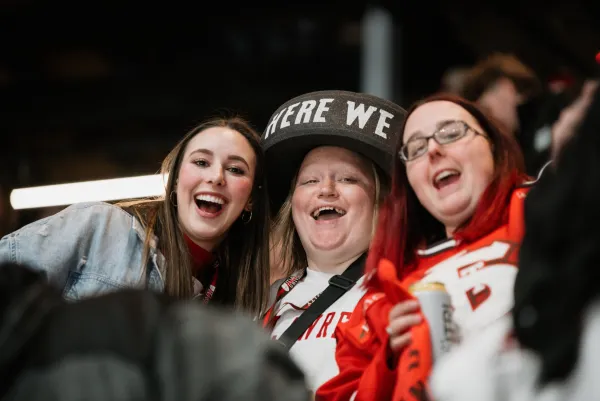 Three young woman stand closely together cheering in the stands of a hockey arena. The woman are wearing hockey jerseys and accessories. One is wearing a black foam hat with white lettering