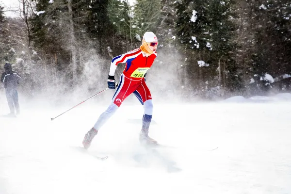 Nordic skier in red race outfit skis through lose snow, kicking up a cloud of snow around them. They wear a yellow race bib on their chest