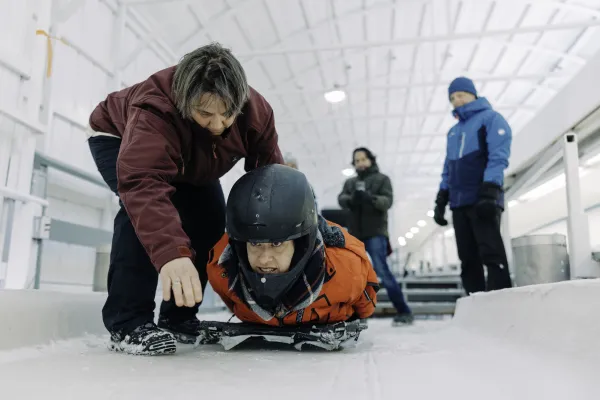 Woman indicates to ice in front of person laying in helmet on skeleton sled on ice track while two other people look on