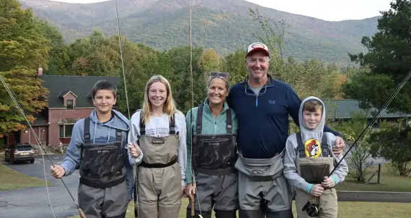 A family of five stands outdoors in waders and holds fishing rods, with mountains in the background.