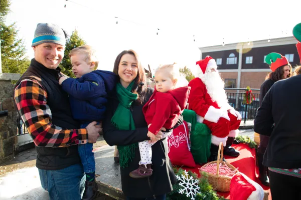 Parents with holding to small children stand outdoors near a Santa Claus seated behind them