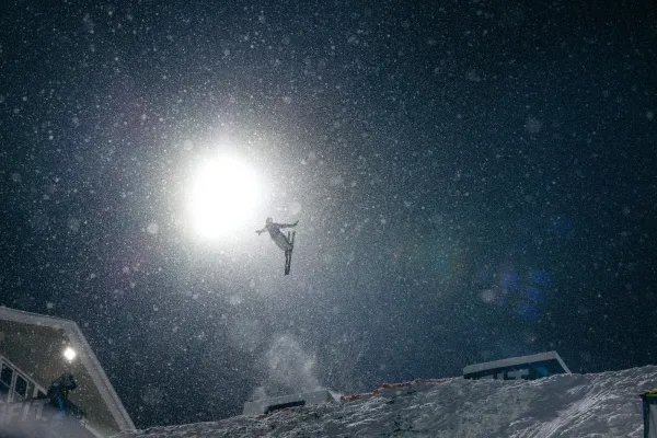 Freestyle aerial skier performs a flip in the air on a dark night with a bright florescent light lighting the skier, ski hill, and snowflakes in the air