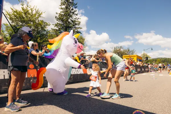 Young girl in white dress accompanied by woman in tshirt and shorts gives a high five to a unicorn mascot while race volunteer waits with finisher medal