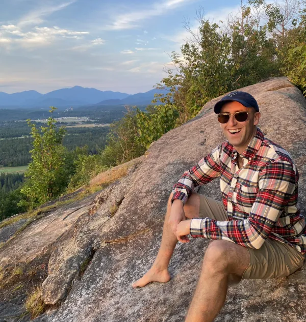 A bare foot man in sunglasses and a flannel sits atop a rocky mountain top.