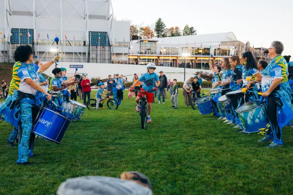 Boy rides bike between rows of a performing drum line as other kids wait their turn and adults look on at Olympic Oval