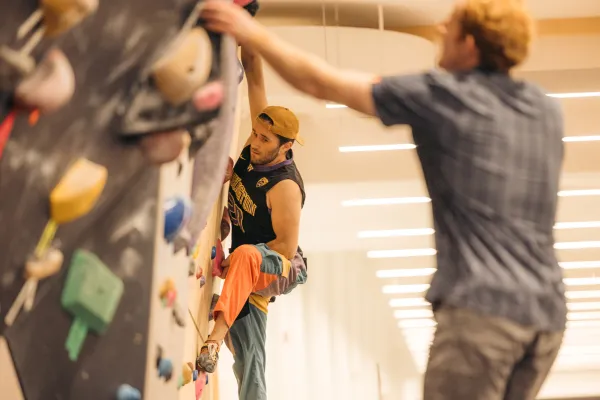 Boulderer seen climbing on indoor rock wall while another person reaching out to touch hold on wall in foreground