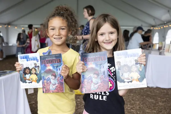 Two young girls stand side by side holding up two children's books each while smiling at the camera