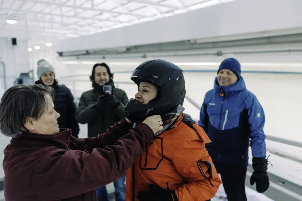 Track worker helps woman put on helmet outdoors at an ice sliding track while others look on