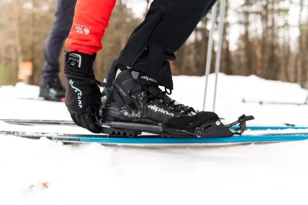 A person in red and black ski gear adjusts their boot binding on a blue ski against a snowy backdrop, conveying a sense of readiness and focus.