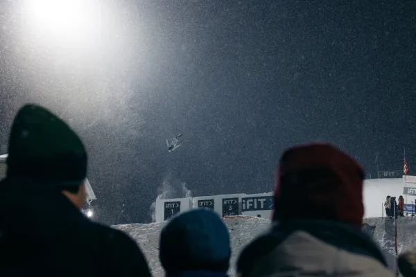 Freestyle aerial skier launches into a flip off a jump on a snowy hill at night, illuminated by artificial light. Dark heads look on in the foreground