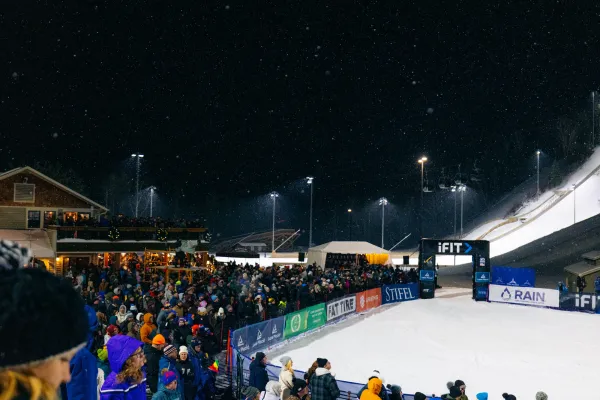 Crowd is seen in front of the Intervale Lodge at the Olympic Ski Jumps for a nighttime freestyle aerials competition. The crowd gathers around the skiers finish area, held back by a barricade.
