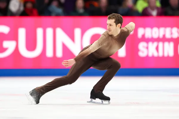 Male figure skater in brown long sleeve and long pant spandex suit performs a crouched move on ice