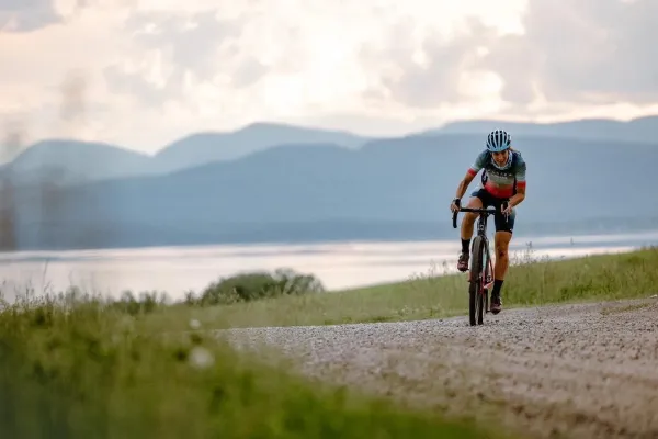 Lea Davison rides mountain bike on gravel road next to large body of water with mountains in the distance