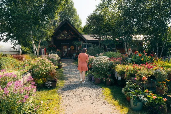 A woman in a pink dress walks down a path lined with blooming flowers toward a wooden building surrounded by lush trees.