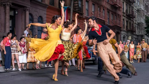 A group of dancers perform a routine on a city street lined, led by a woman in a yellow dress and a man in a polo shirt.