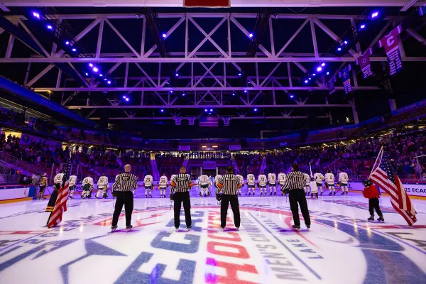 Indoor ice hockey arena filled with spectators. Rink is well-lit, featuring a central logo and teams preparing for a face-off. 