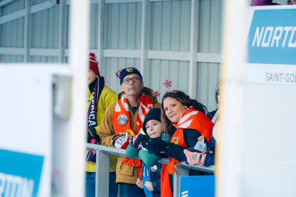 Spectators wearing winter and country-specific attire stand under awning looking onto luge track