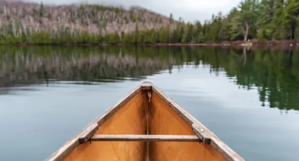 Wooden canoe front on placid lake with evergreen trees and leafless deciduous trees on the shoreline