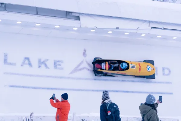 Yellow bobsled descends ice track with Lake Placid logo frozen into ice while people take photos and look on