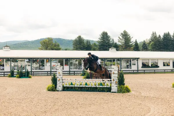 Equestrian rider jumps horse over jump on dirt ring with pavilion building in background