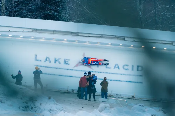View partially obscured by trees of luge athlete riding down track over where Lake Placid is printed into the ice while several people look on