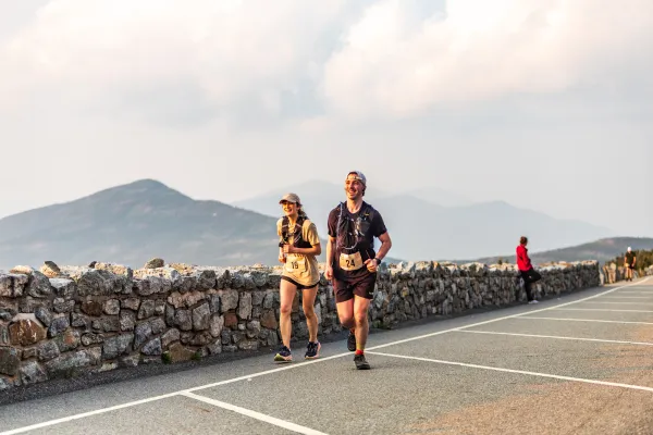 Two smiling runners wearing race bibs and hydration vests jog up a mountain road lined by a stone wall.