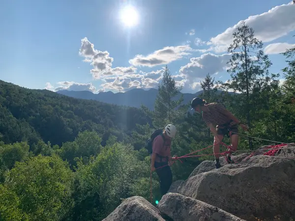 Two rock climbers on a sunny day, one belaying the other on a rocky outcrop overlooking a lush green valley and distant mountains.