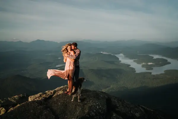 A couple embraces on a rocky ledge while her dress and hair flow in the breeze with Lake Placid in the back drop