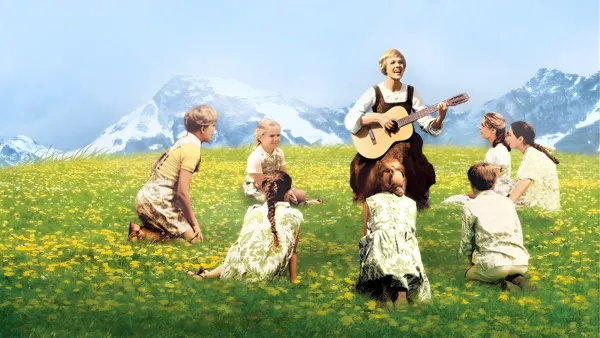 Maria from *The Sound of Music* sits in a sunlit, wildflower meadow playing a guitar and singing to children around her.