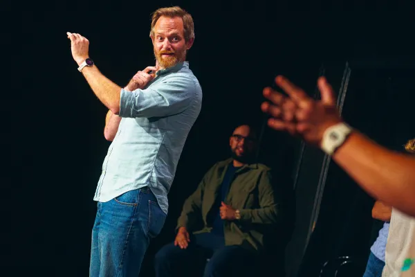 Bearded man in dress shirt and jeans holds up one hand and looks and points in the direction of someone out of image adjacent