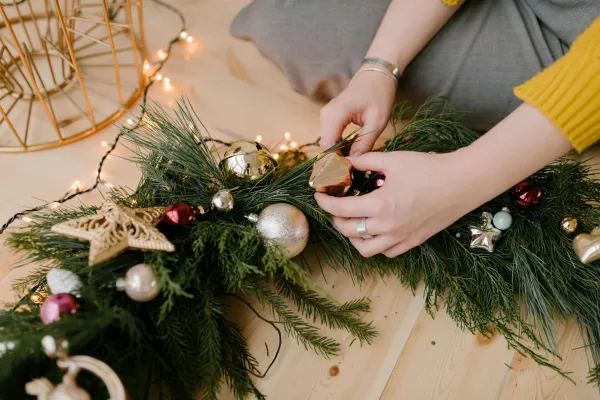 A close-up shot of a person arranging ornaments and pine sprigs to create a Christmas garland on a light wooden floor, surrounded by twinkling string lights.