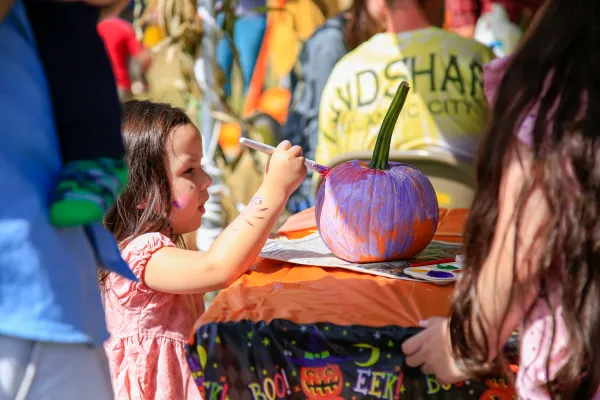 Young brunette girl in a pink dress paints a small pumpkin at a fall event