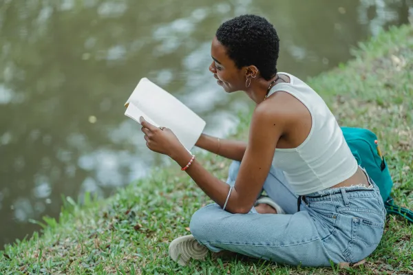 Young woman sits next to lake on grass reading a book wearing jeans and white tank top