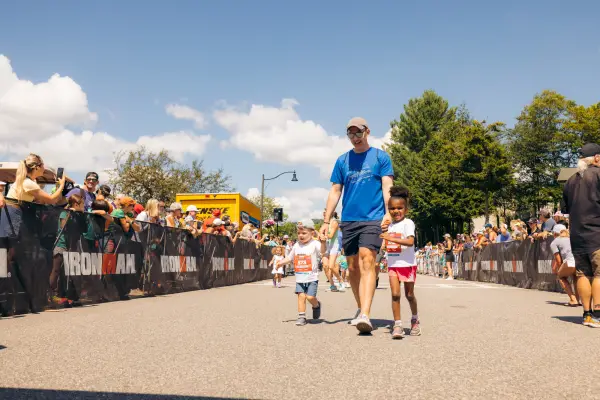 Man in blue tshirt and shorts wearing hat and sunglasses walks with two young children, each holding one of the man's hands, on a race course lined by spectators and Ironman race fencing