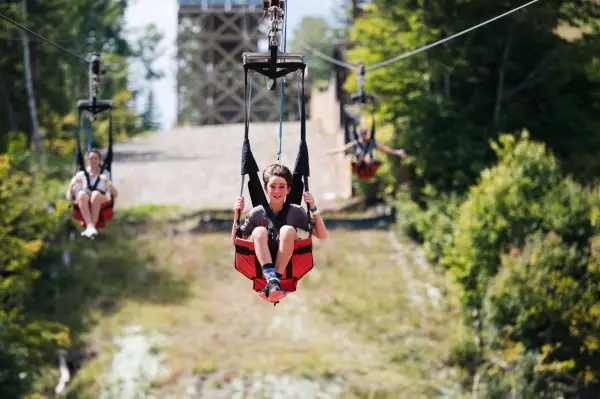 Young boy sits in sling seat of zipline smiling with wind blowing back his hair as two other youths follow behind