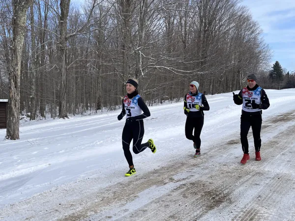 Three runners in winter gear race on a snowy, tree-lined path. The lead runner smiles, conveying determination and joy.