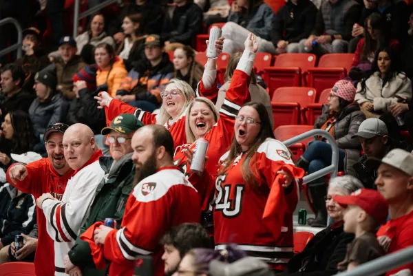 A group of three women in red hockey jerseys cheer and look toward the camera in the crowded stands of the 1980 Herb Brooks Arena during an ECAC Hockey game. 