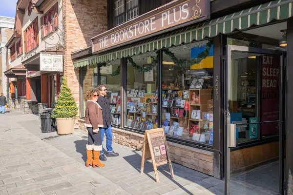 Man in black peacoat and woman in brown leather coat stand outside The Bookstore Plus on Main St Lake Placid