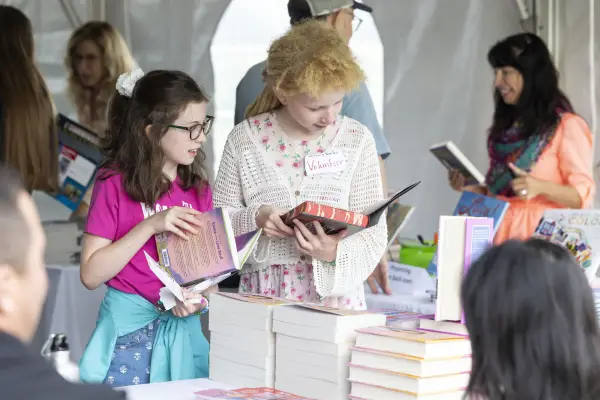 Two girls look at books in front of table stocked with tall piles of books