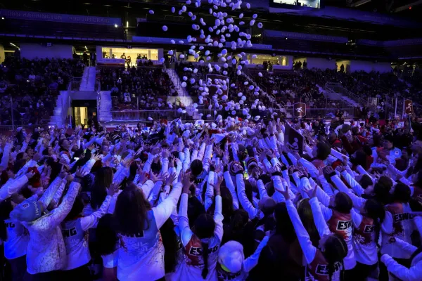 Crowd of people in a dimly lit arena reaching up as white "snowballs" rain down. Excitement fills the scene, with spectators in colorful team uniforms.