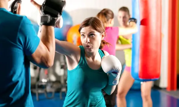A woman practicing throwing a punch in a boxing ring with two other women using a punching bag in the background