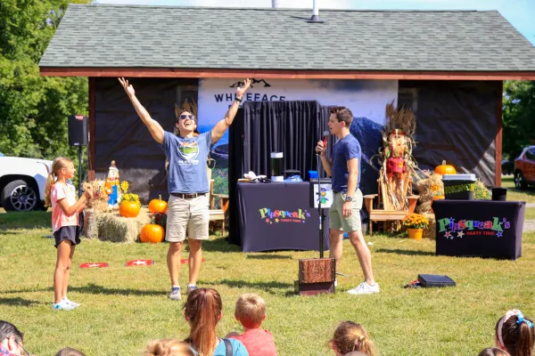 Two men stand at front of crowd of children sitting on grass at fall event, one talking on a microphone and the other raising both arms