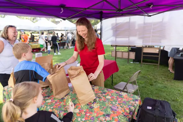A woman at a farmer's market fills brown paper bags with produce for children's activities.