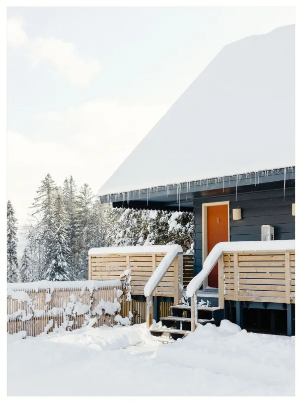 The exterior of one of the cabins with large porch blanketed in snow with tall pines in the background.