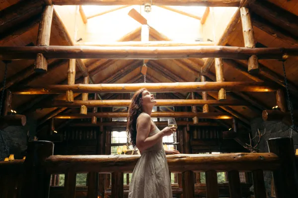 A woman stands on the balcony of a grand lodge with a glass of wine in her hand.