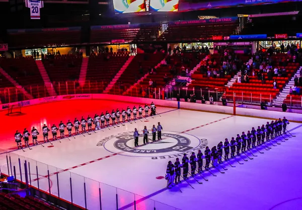 Two teams of female hockey players lined up opposite one another on the ice with officials in the middle under colorful light