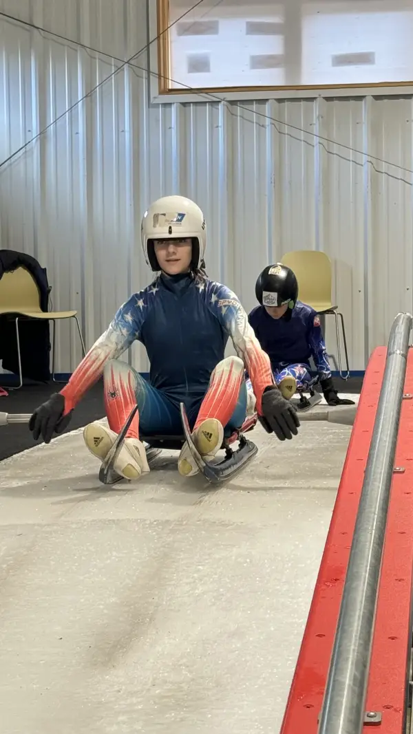 A young woman in a USA luge uniform sits on a luge sled on an indoor track.  Another young person is visible behind her, also on a sled.