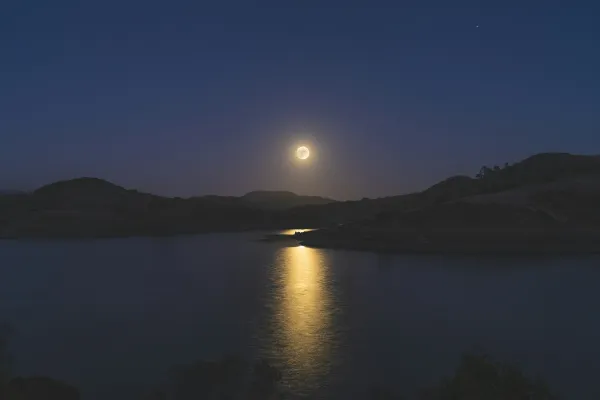 Full moon in dark sky over lake and trees shoreline hills with moon reflecting in the water