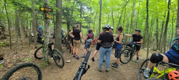 A group of women mountain bikers pause on a trail in a wooded area to chat.  Several bicycles are parked nearby.