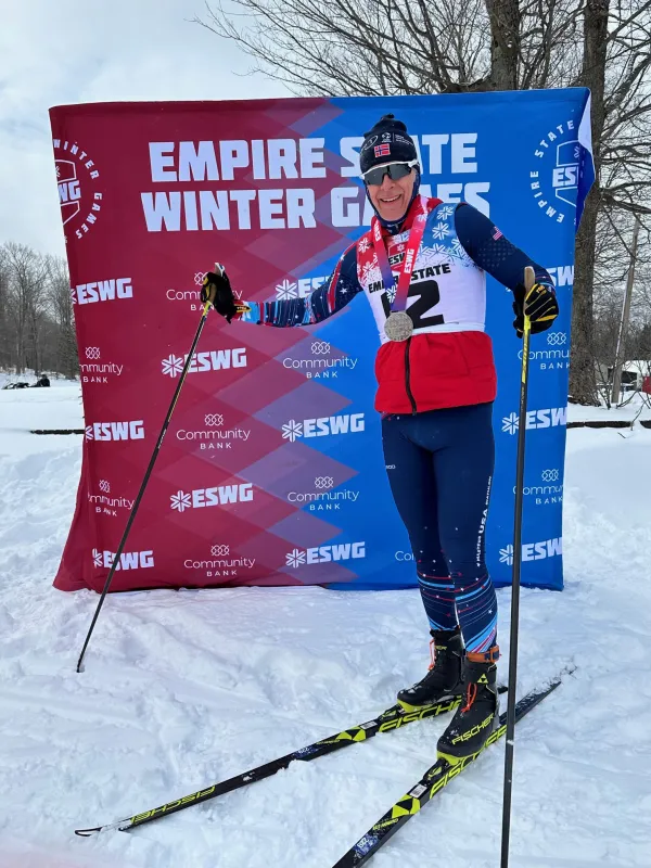 Skier with medals and ski poles poses proudly in front of a branded Empire State Winter Games backdrop, wearing a red vest and blue tights on snow.